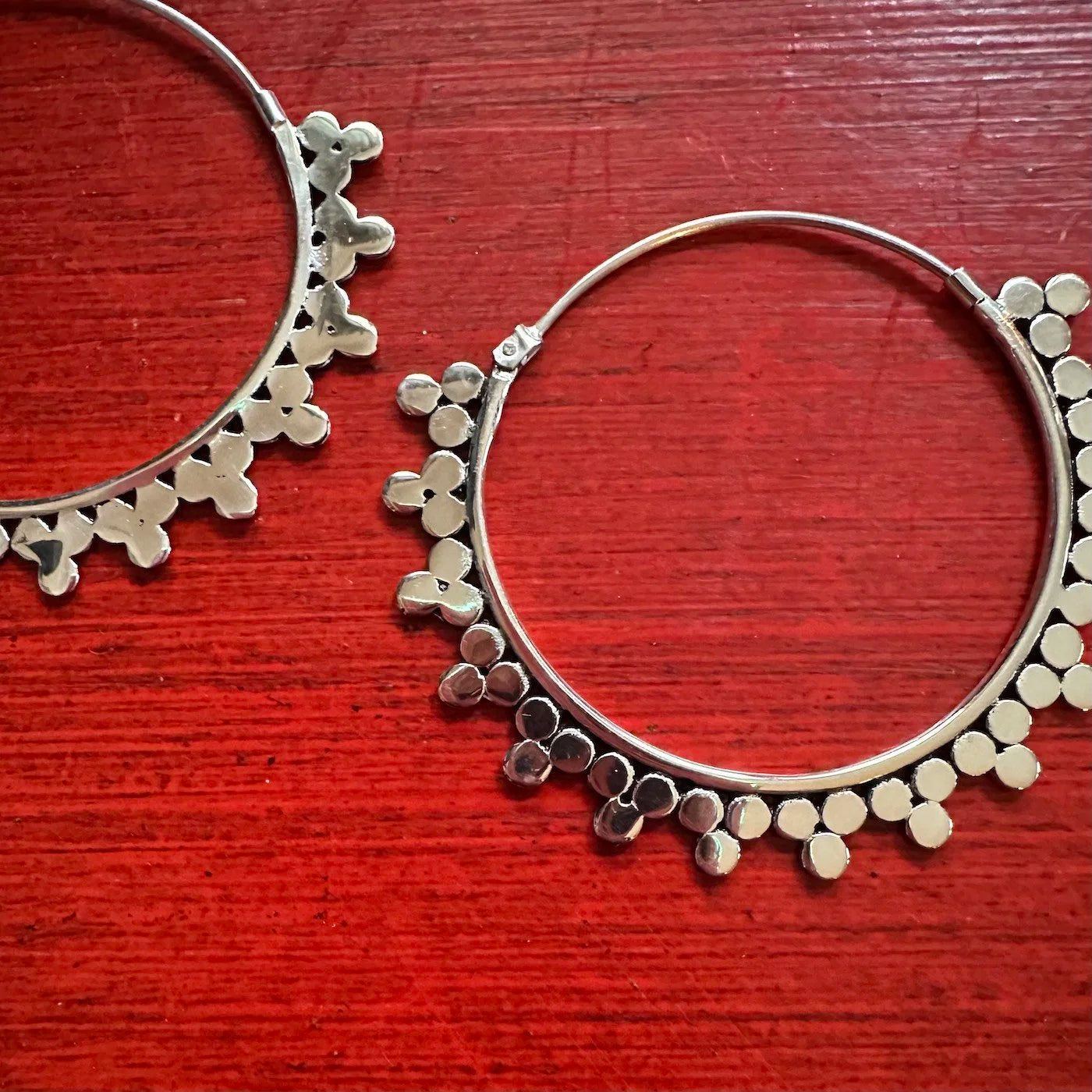Silver Indonesian hoop earrings with floral and dot patterns on a red wooden surface