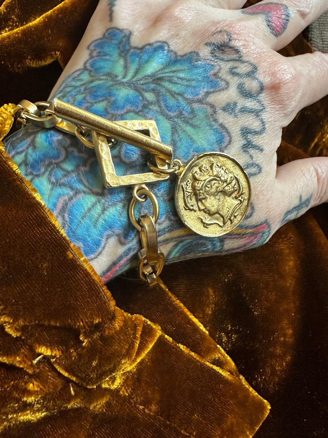 Gold bracelet with a Roman coin charm on a hand with tattoos against a brown velvet background