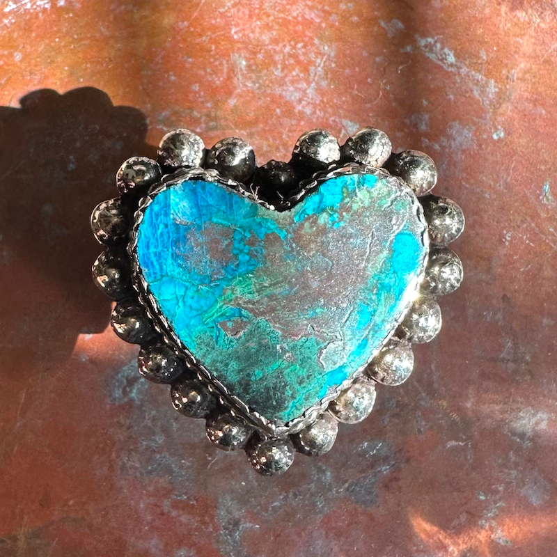 Heart-shaped chrysocolla turquoise stone ring with silver border on a textured surface